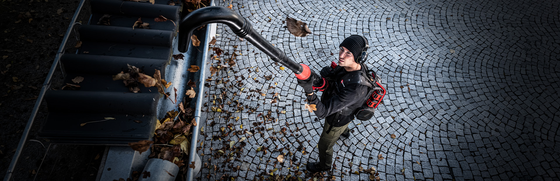 A man uses an Einhell cordless leaf blower with a long gutter cleaning attachment to clear leaves from a gutter.