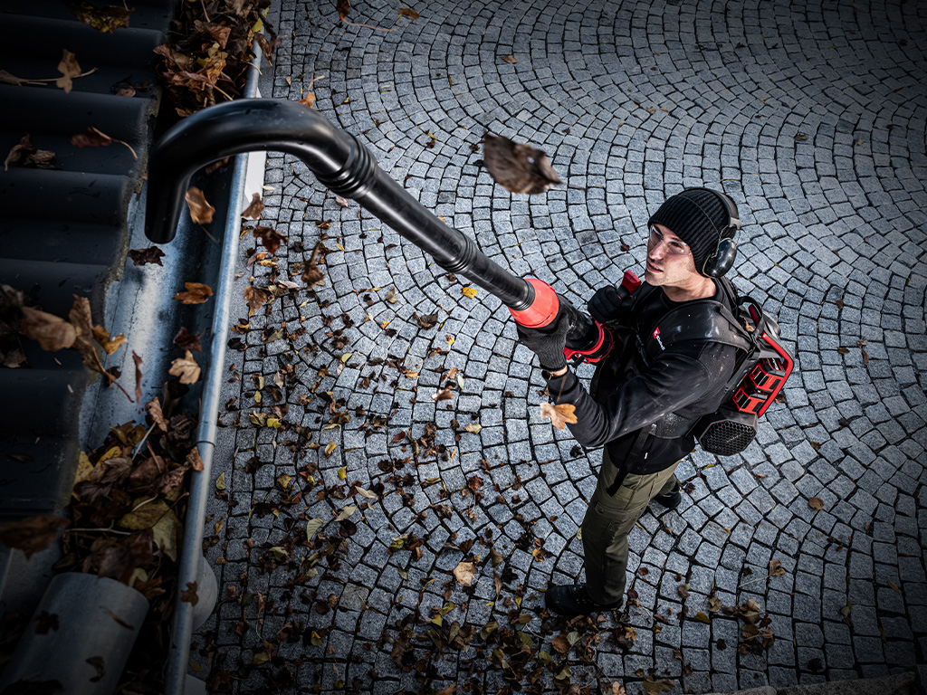 A man uses an Einhell cordless leaf blower with a long gutter cleaning attachment to clear leaves from a gutter.