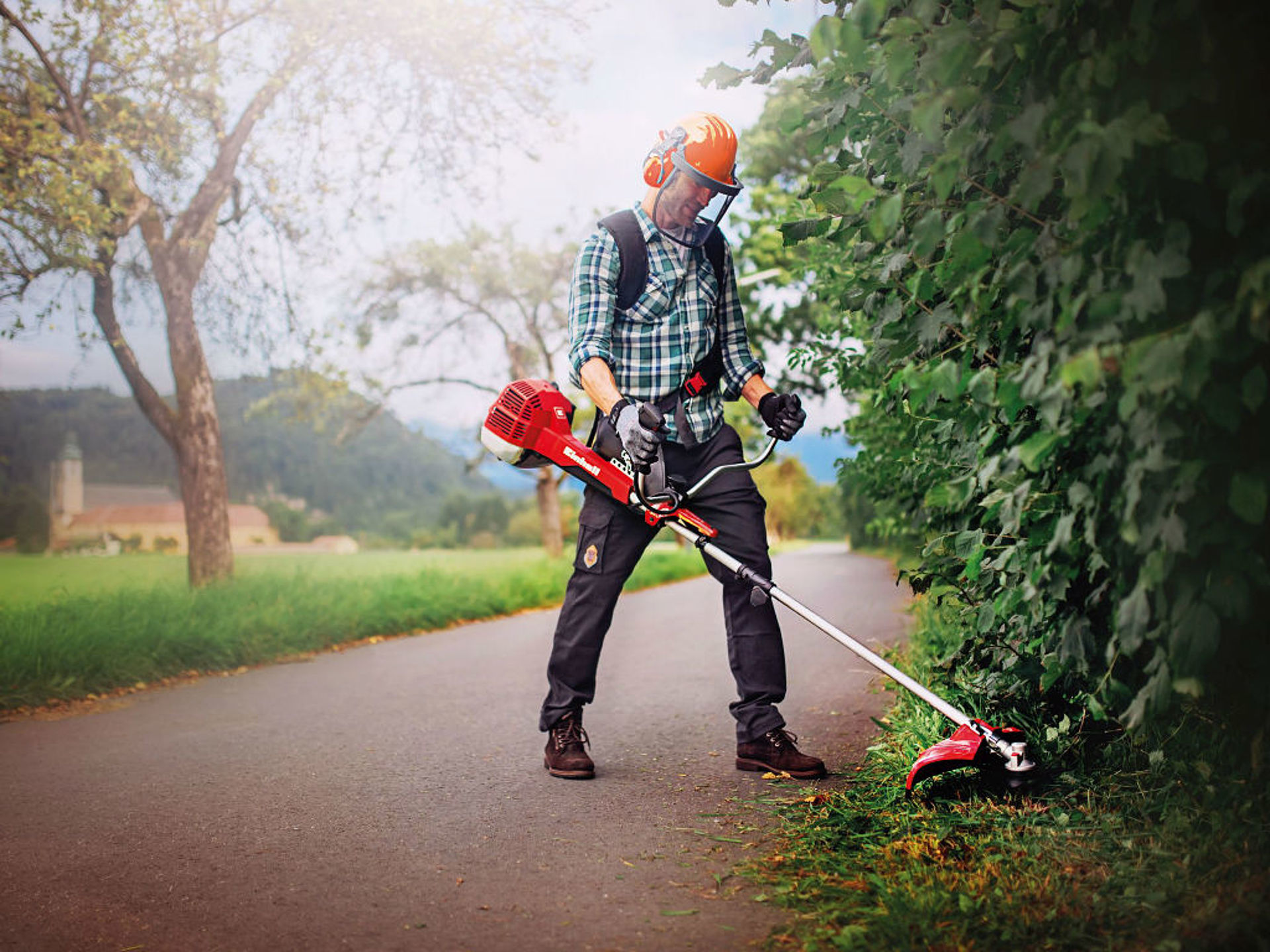 A man cuts grass under a hedge with a cordless scythe from Einhell.