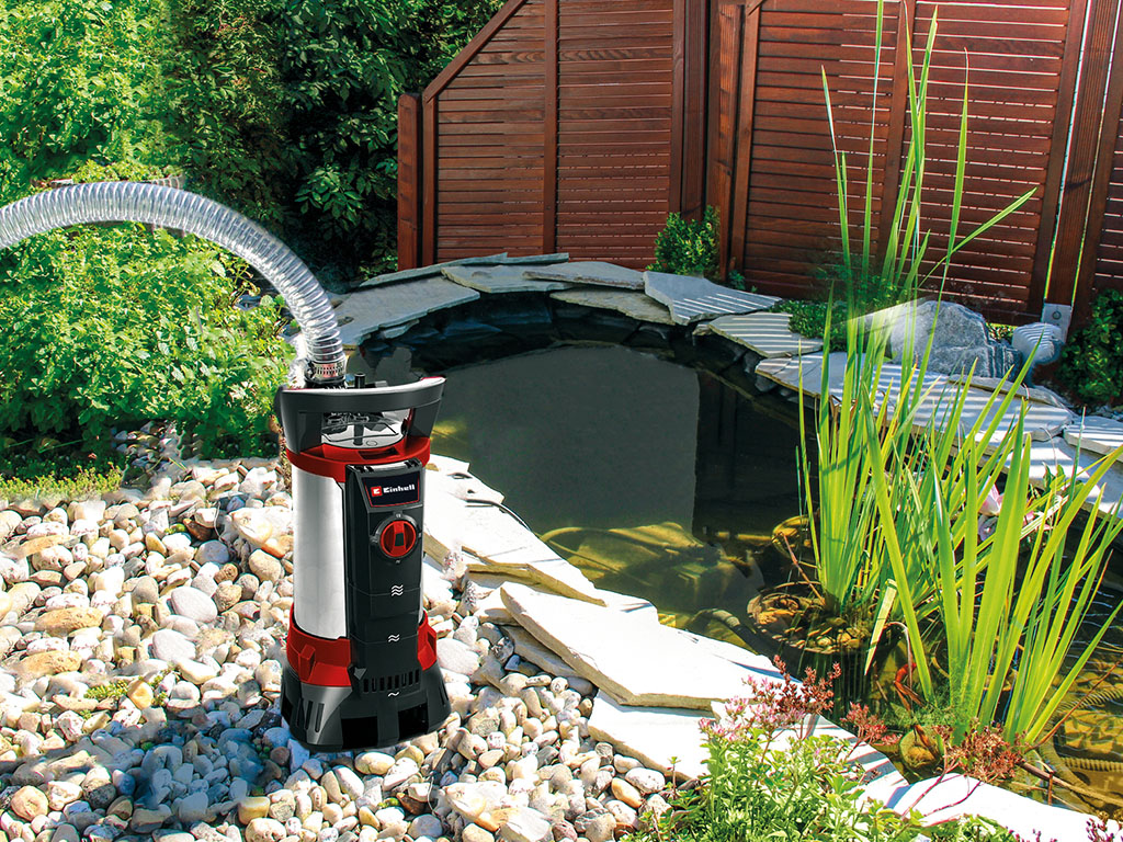 Eine Einhell-Schmutzwasserpumpe steht neben einem Gartenteich mit Steinen, Pflanzen und kleinem Wasserfall.