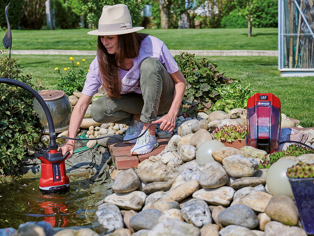 Eine Frau benutzt eine Einhell-Schmutzwasserpumpe, um Wasser aus einem Gartenteich neben einem Steinhaufen abzusaugen.