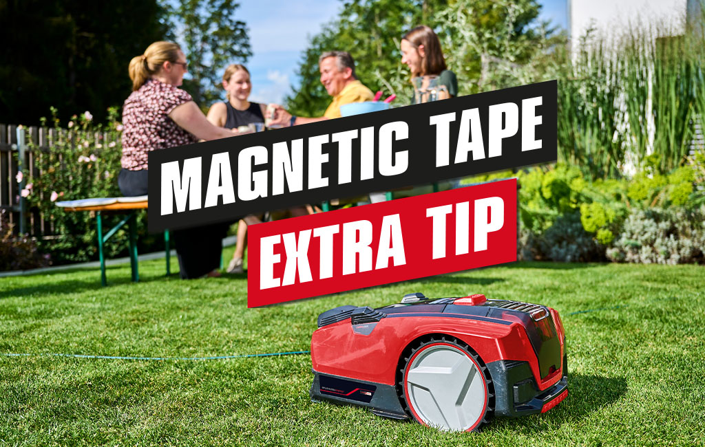 A robot lawnmower mows the lawn in a garden where four people are sitting at a table. The text in the picture reads: magnetic tape - extra tip.
