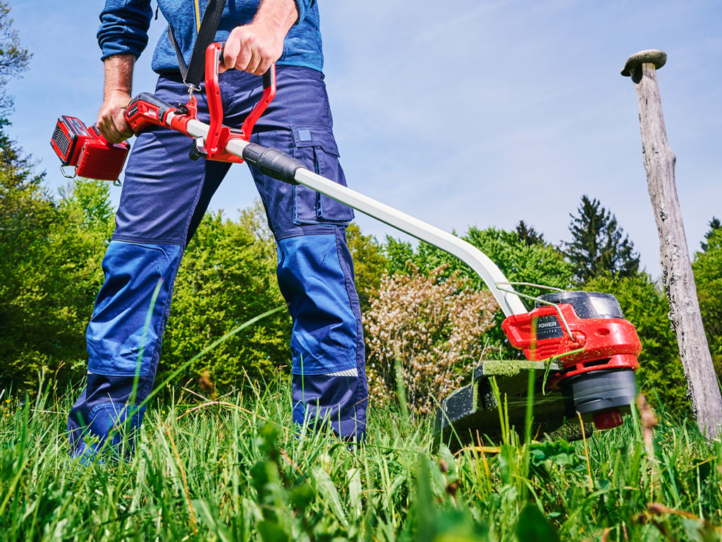 Ein Mann mit blauer Hose und rotem Akku-Rasentrimmer trimmt das Gras auf einer Wiese.