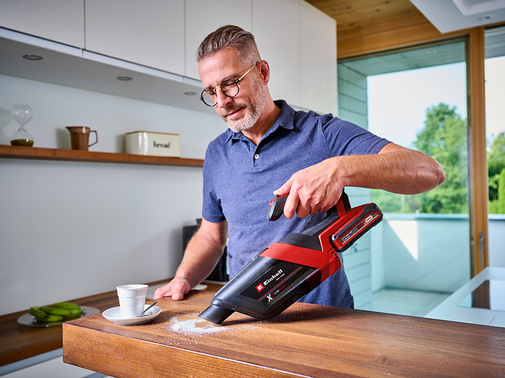 Ein Mann saugt mit einem roten kabellosen Handstaubsauger Krümel von einer Küchenarbeitsplatte.
