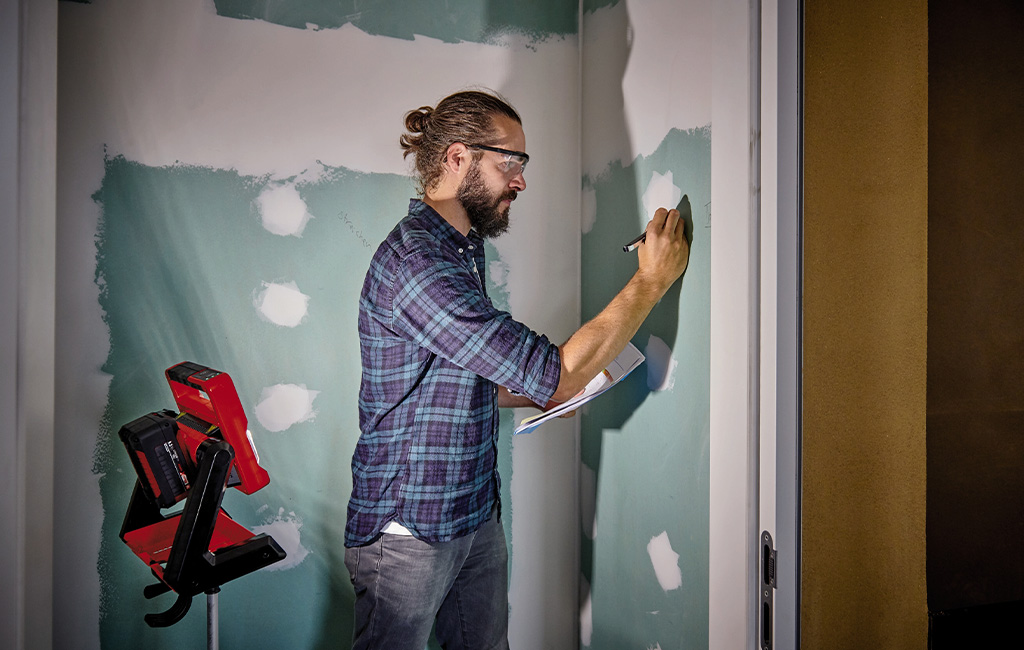 A man drawing with a pencil on a piece of drywall illuminated by a battery-powered lamp.