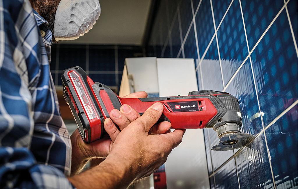 A craftsman cuts the grout of dark wall tiles using an Einhell cordless multitool.
