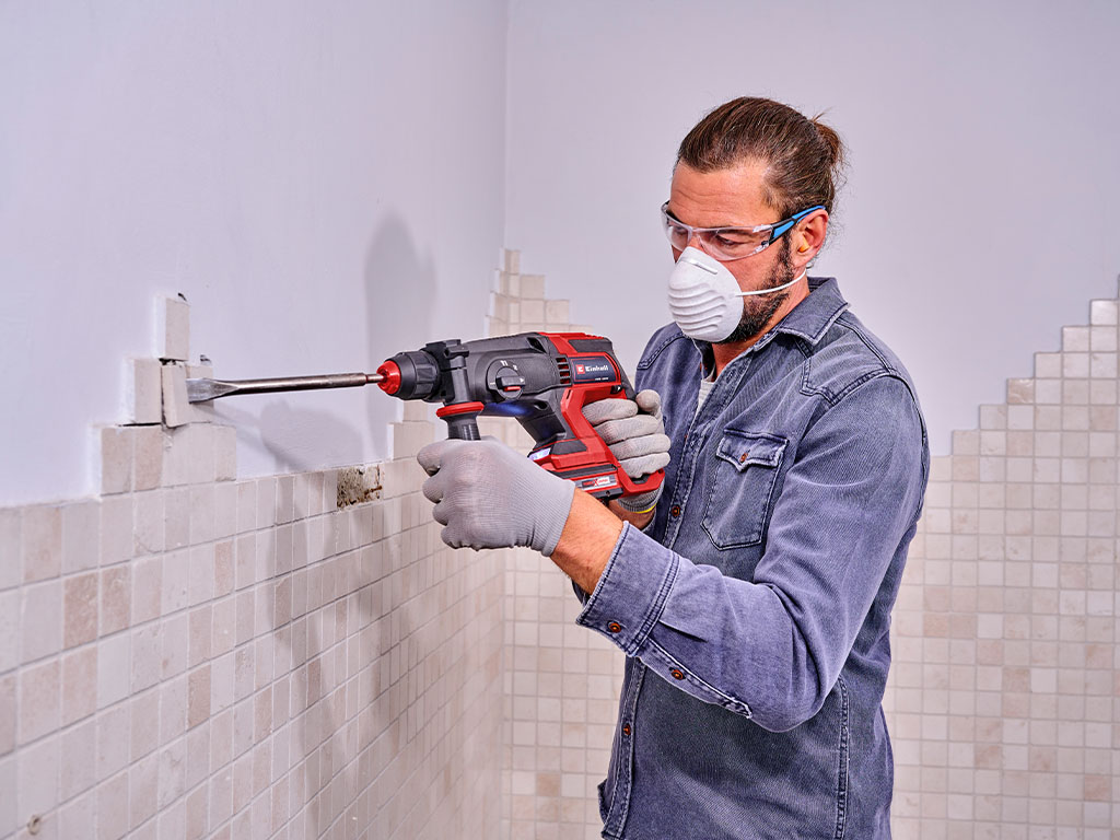 A man wearing safety glasses, gloves, and a dust mask removes wall tiles using an Einhell cordless rotary hammer.