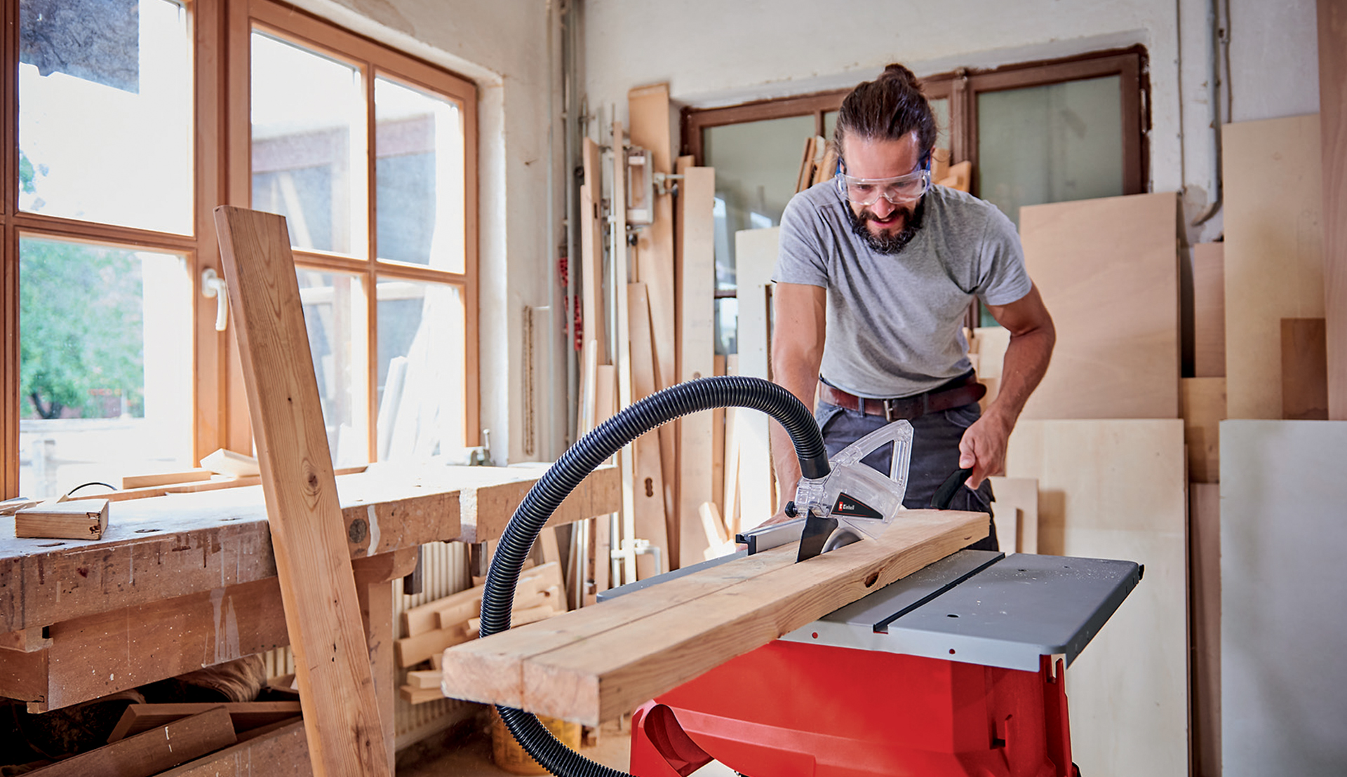 A man guiding a wooden board across a table saw with a dust extraction hood in a workshop.