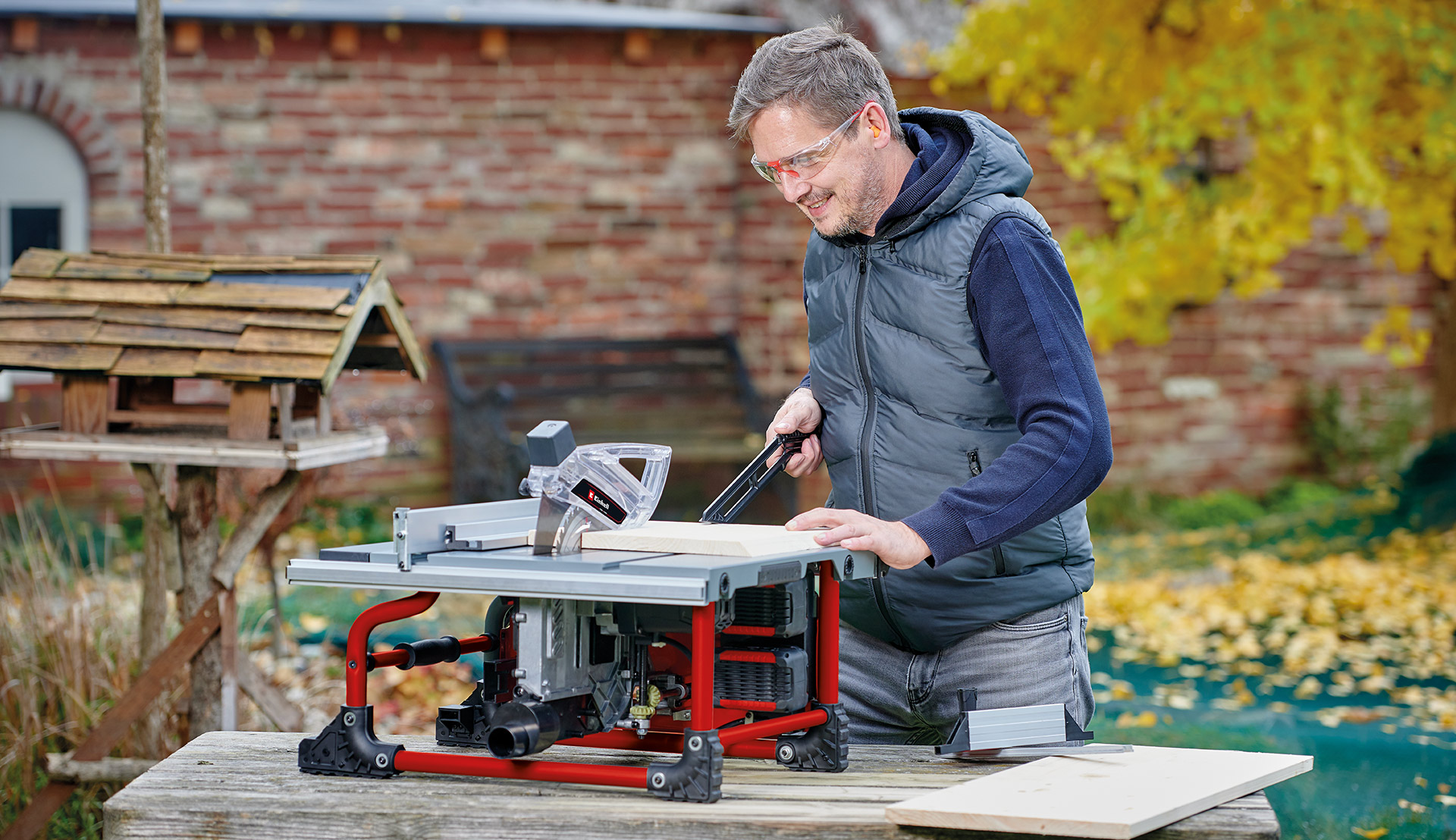 A man working outdoors with a table saw, guiding a wooden board across the saw table.