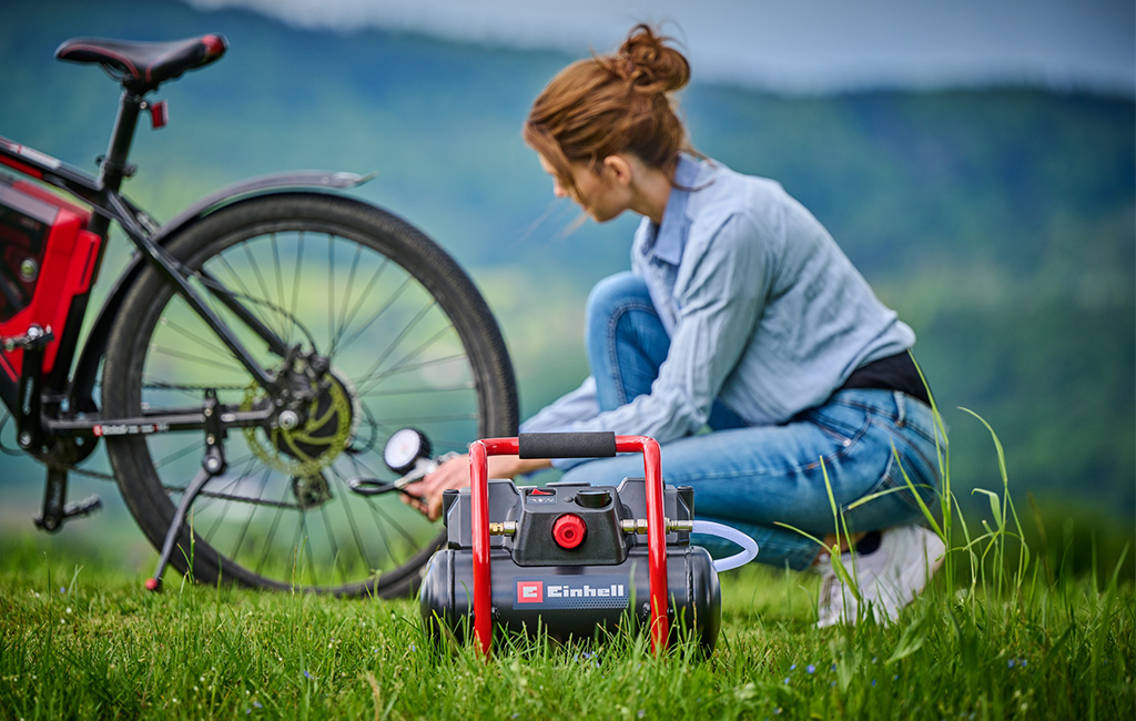 Eine Frau pumpt mit einem Einhell Akku-Kompressor den Reifen eines Fahrrads auf, während sie in einem grünen Feld sitzt.
