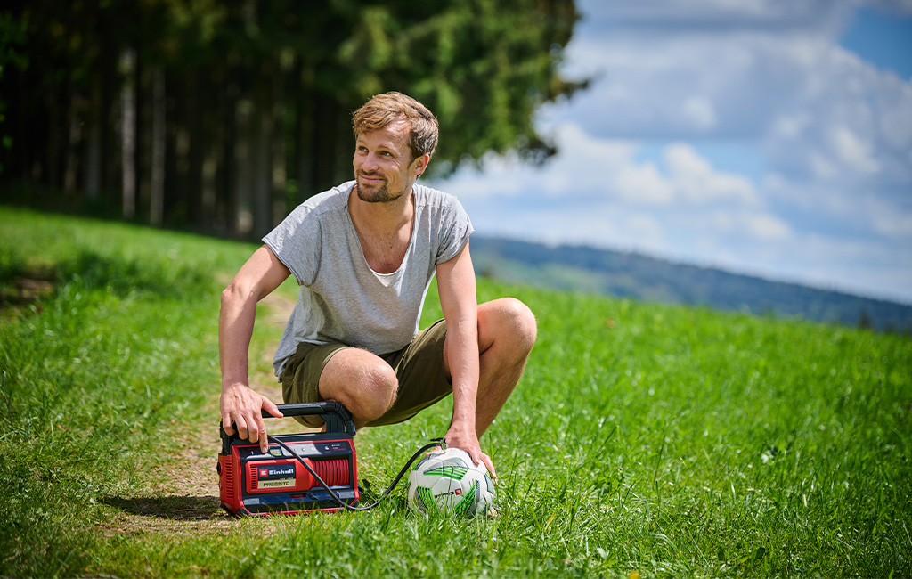 Ein Mann kniet auf einer Wiese und nutzt den Einhell Akku-Kompressor PRESSITO, um einen Fußball aufzupumpen.