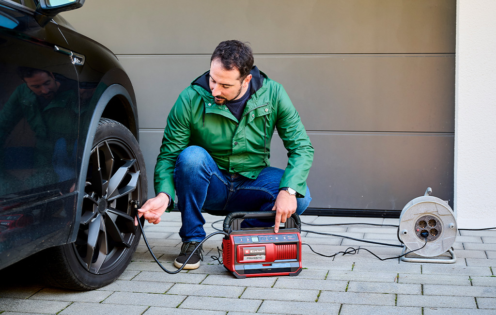 Ein Mann pumpt mit dem Einhell Hypbrid-Kompressor PRESSITO den Reifen eines Autos auf, während er vor einer Garage kniet.