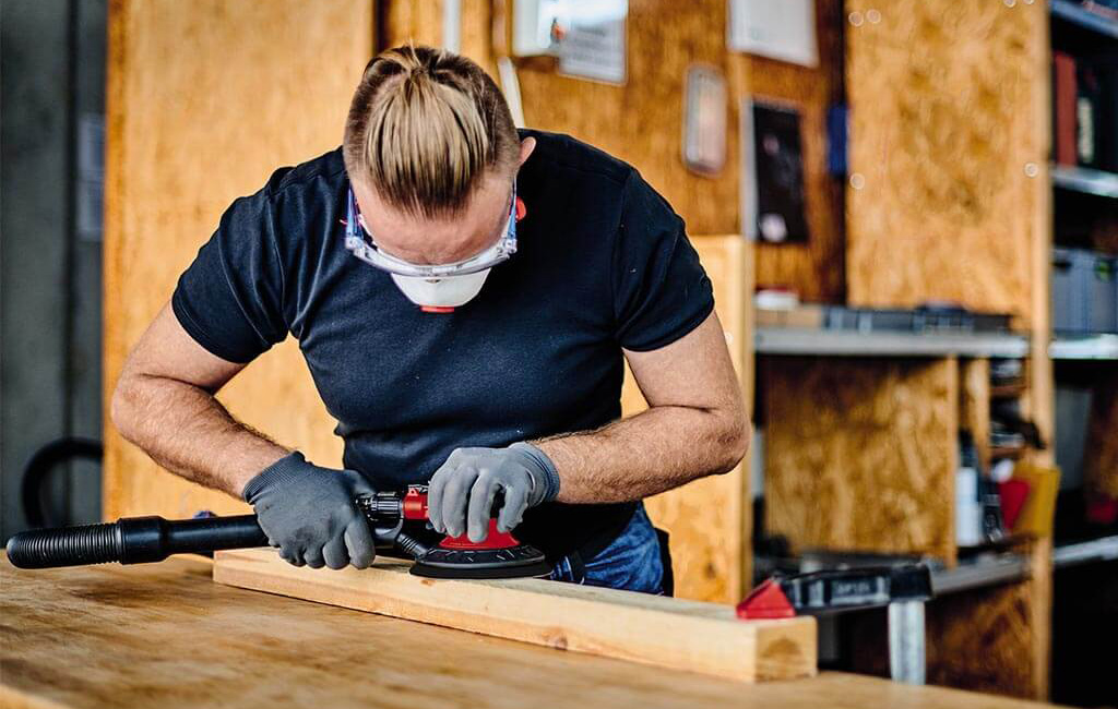 Ein Mann schleift ein Holzbrett mit einem Druckluft-Schleifwerkzeug in einer Werkstatt.