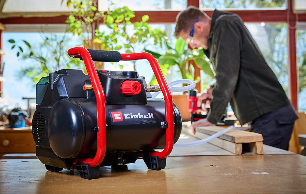 Ein Mann arbeitet in einer Werkstatt mit einem mobilen Einhell Akku-Kompressor auf einem Tisch im Vordergrund.