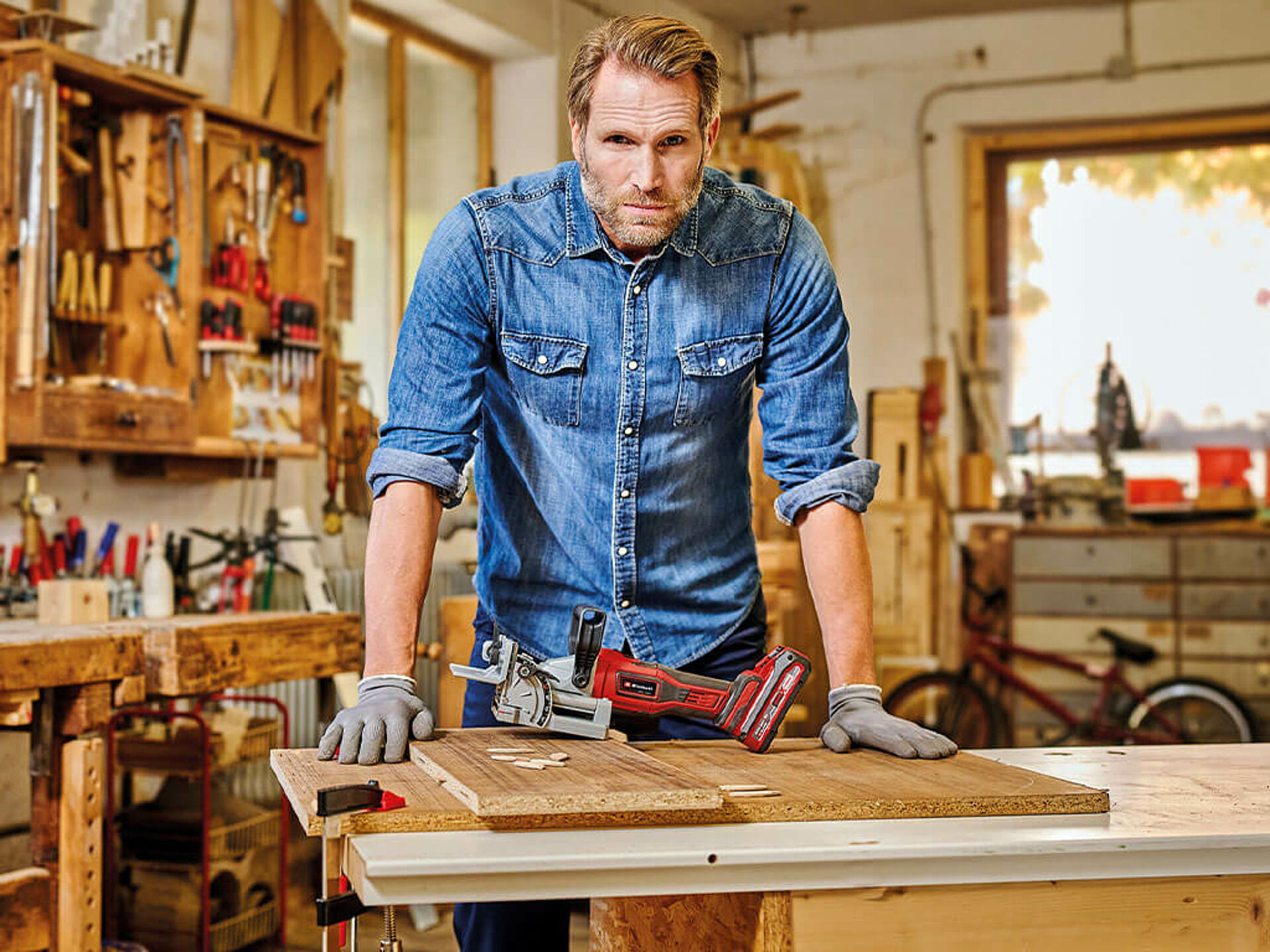 Ein Mann lehnt sich auf eine Werkbank mit der Einhell Akku-Flachdübelfräse, Dübeln und Holzplatten. Er blickt ernst in die Kamera.