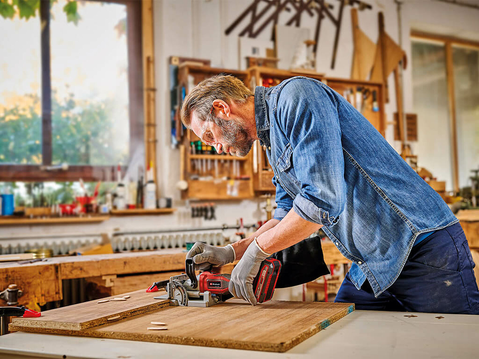 Ein Mann arbeitet mit der Einhell Akku-Flachdübelfräse in einer Werkstatt an zwei Holzplatten. Dübel und Schraubzwinge liegen bereit.