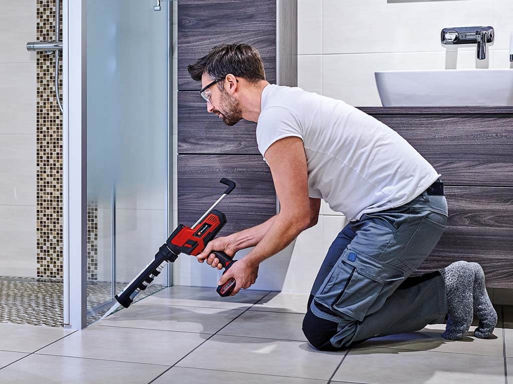 A man kneeling in the bathroom and applying silicone between the shower wall and the tiled floor.