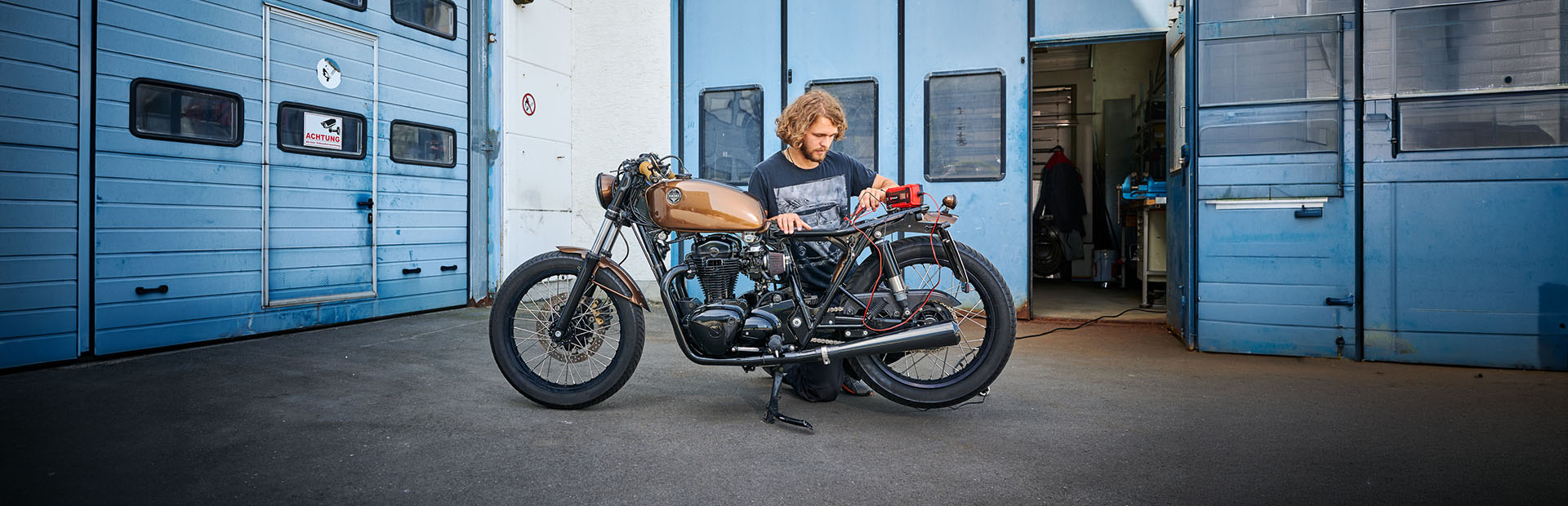 Person kneeling next to a motorcycle in front of a workshop checking the electrics with a measuring device.