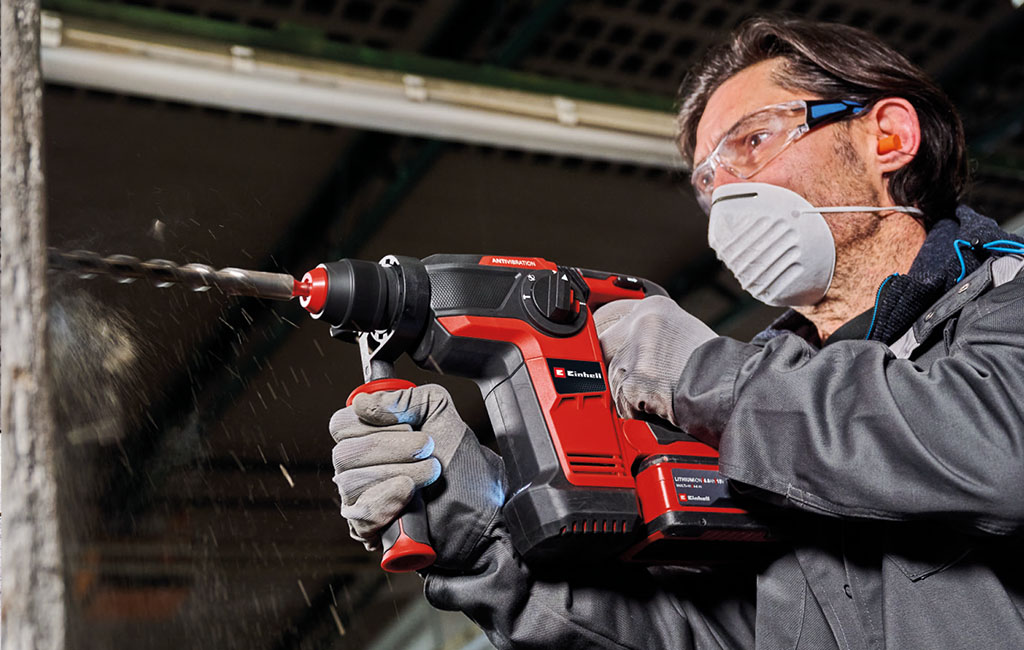Man drilling into a concrete wall with a cordless rotary hammer while wearing safety glasses, a dust mask and hearing protection.