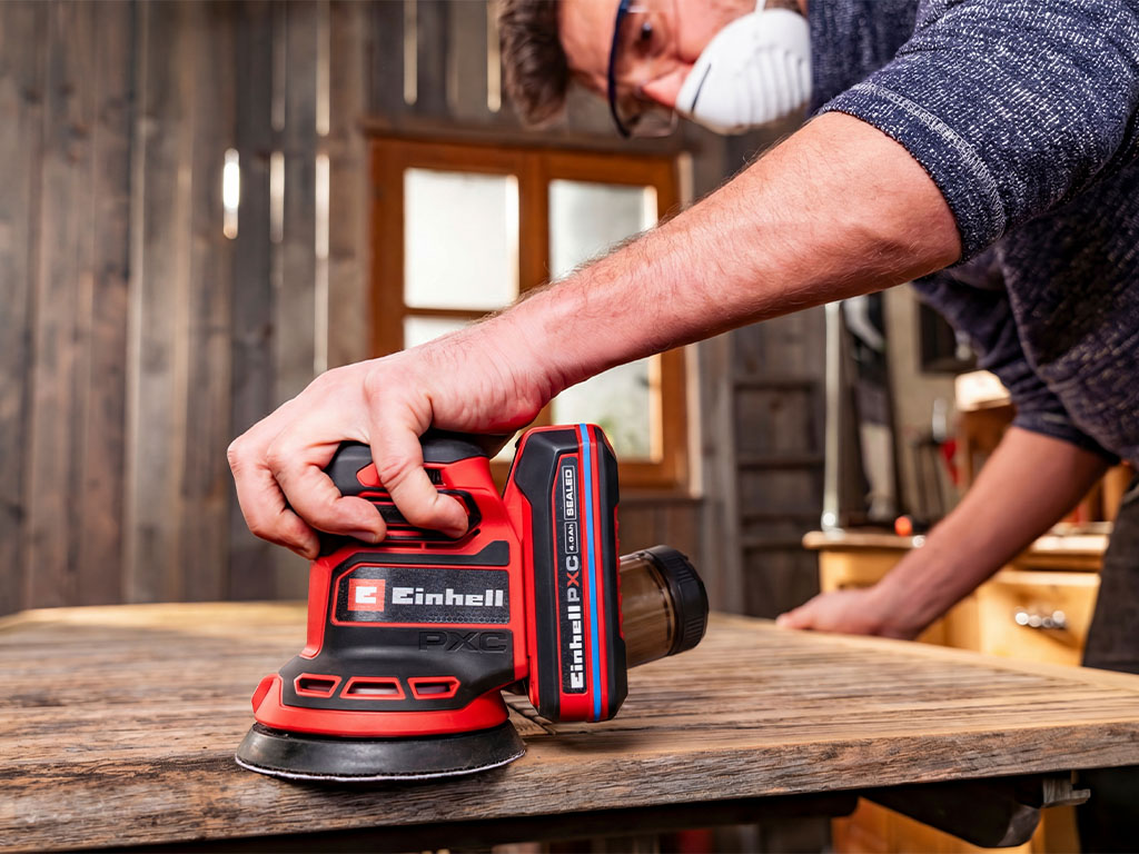 A man works on a wooden surface in a garage using a random orbital sander.