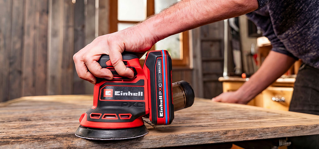 A man works on a wooden surface in a garage using a random orbital sander.