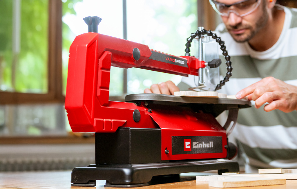 Ein Mann arbeitet mit einer roten Einhell Dekupiersäge an einem Holzstück auf einem Tisch.