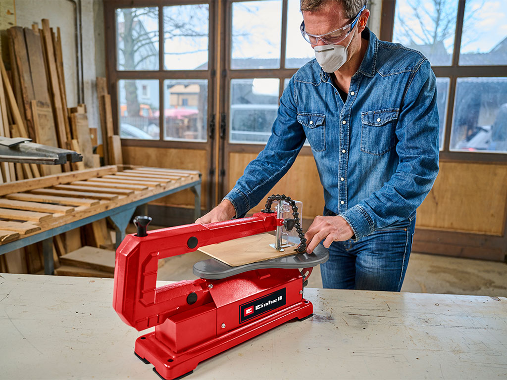 A man stands at a table and works on a wooden board using the red Einhell scroll saw.