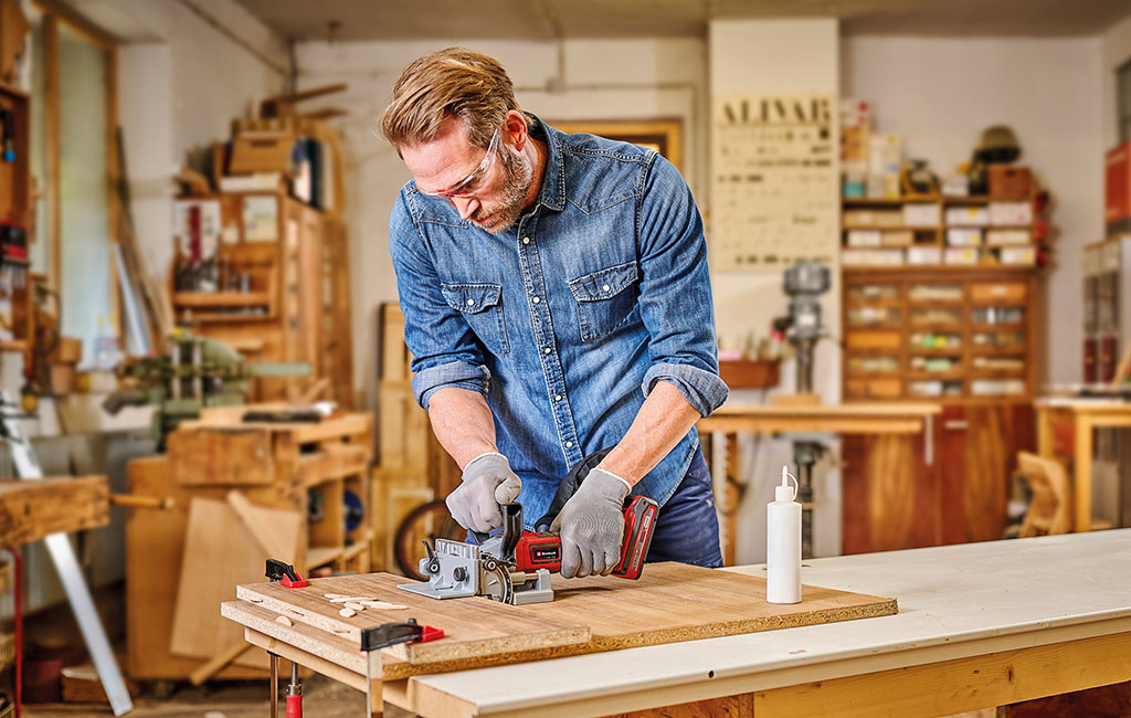 Ein Mann fräst in einer Werkstatt mit einer Einhell Akku-Flachdübelfräse Schlitze in Holzbretter für ein DIY-Projekt.