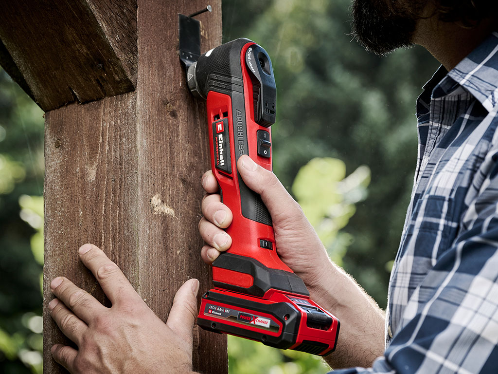 A man holds a red multifunctional tool against a wooden beam, precisely working on the surface.