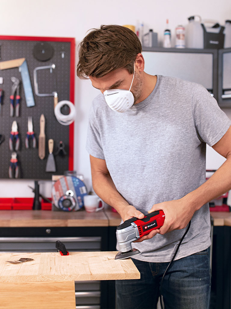 A man holds a red multifunctional tool against a wooden beam, precisely working on the surface.