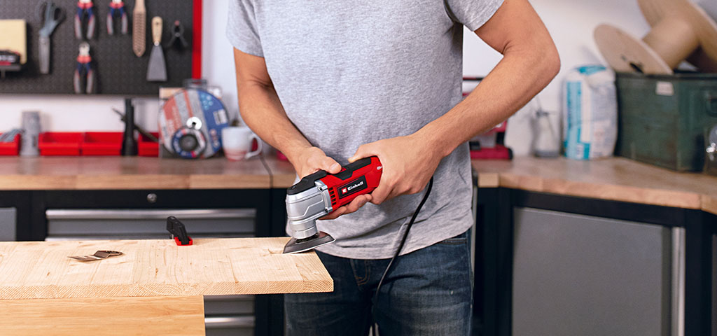 A man holds a red multifunctional tool against a wooden beam, precisely working on the surface.