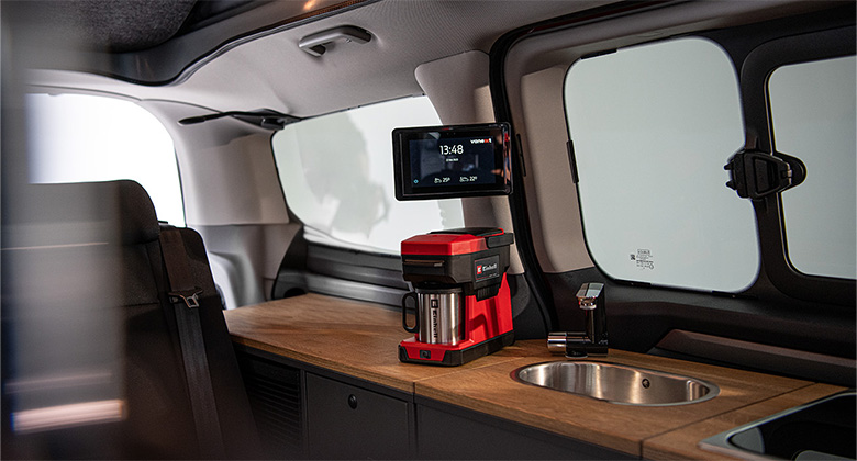 Innenraum eines Camping-Vans mit Arbeitsfläche, Spüle und Einhell-Akku-Kaffeemaschine. Darüber hängt ein kleiner Bildschirm mit der Uhrzeit.