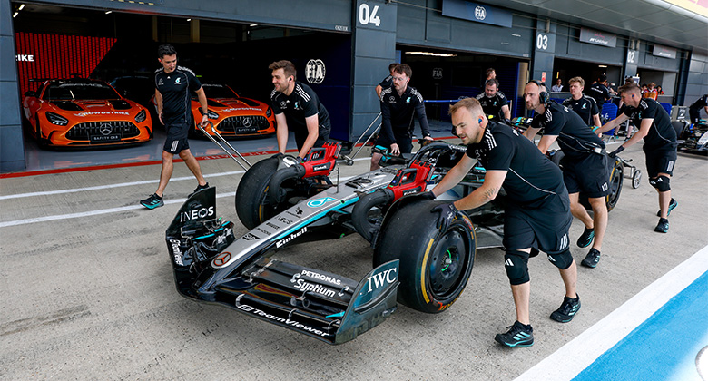 Several team members push a Mercedes-AMG PETRONAS Formula One race car out of the pit lane, with two orange Mercedes-AMG GT cars in the background.