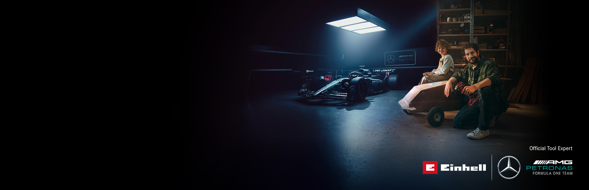 Ein Vater und sein Kind sitzen in einer Werkstatt vor einem selbstgebauten Seifenkistenauto. Im Hintergrund steht ein Mercedes-AMG PETRONAS Formel-1-Wagen mit Einhell-Werkzeugen daneben.