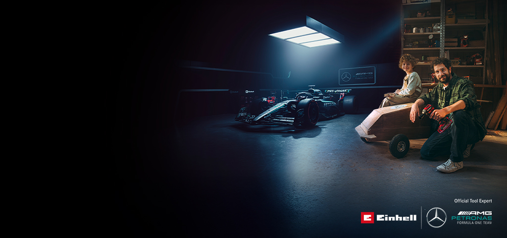 Ein Vater und sein Kind sitzen in einer Werkstatt vor einem selbstgebauten Seifenkistenauto. Im Hintergrund steht ein Mercedes-AMG PETRONAS Formel-1-Wagen mit Einhell-Werkzeugen daneben.