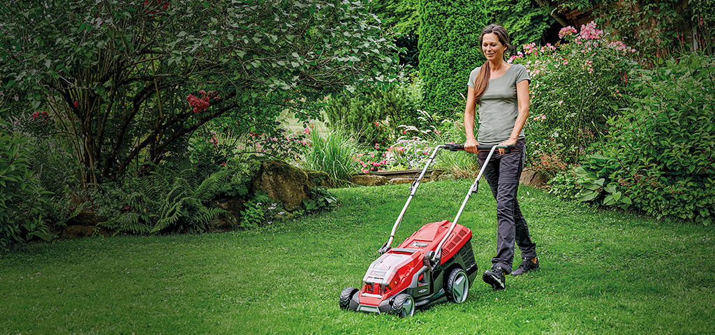 A woman is mowing the lawn in a green, blooming garden with a red Einhell lawn mower.