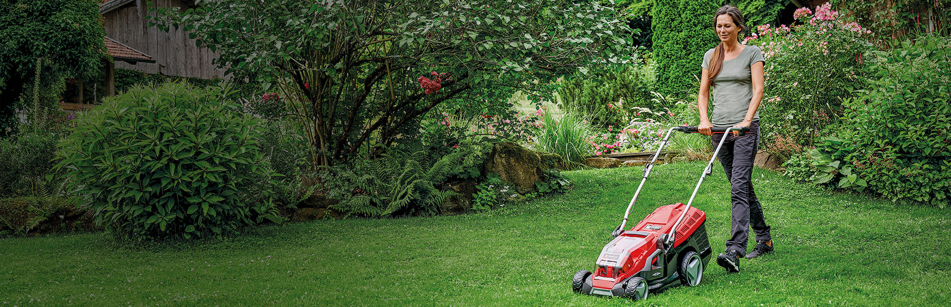 A woman is mowing the lawn in a green, blooming garden with a red Einhell lawn mower.