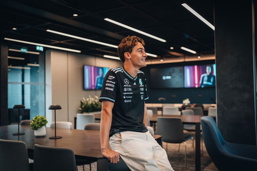 George Russell, Fahrer des Mercedes-AMG PETRONAS Formel-1-Teams, sitzt entspannt auf einem Tisch in einem modernen, stilvollen Hospitality- oder Bürobereich des Teams. Er lächelt und trägt das offizielle Teamshirt mit sichtbaren Sponsorenlogos wie Einhell, IWC und CrowdStrike. Im Hintergrund sind Bildschirme mit seinem Bild zu sehen.
