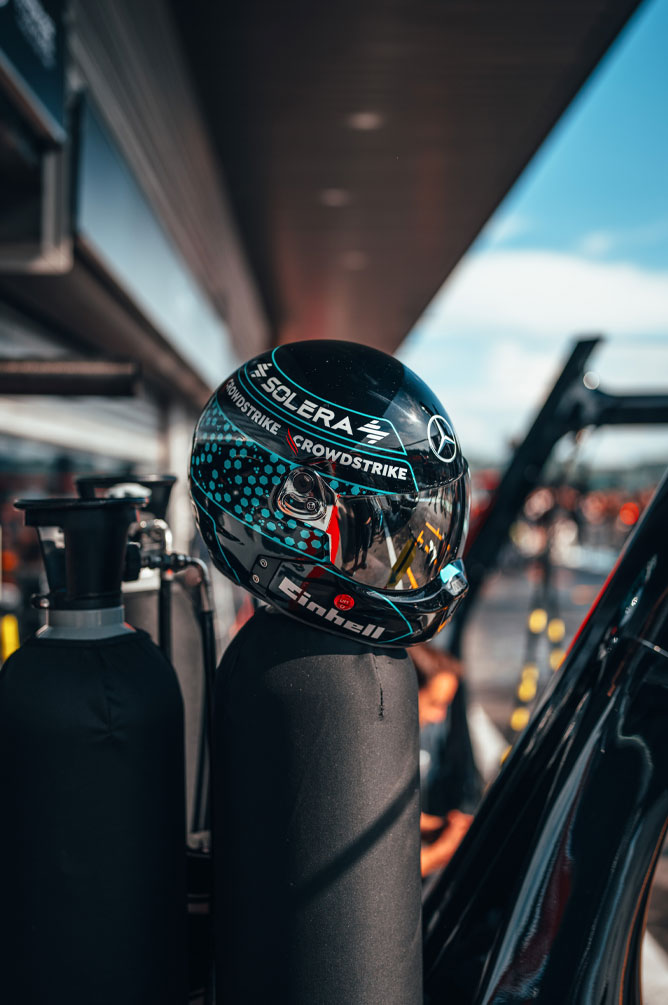 Ein Helm des Mercedes-AMG PETRONAS Formel-1-Teams mit gut sichtbaren Sponsorenlogos wie Einhell, CrowdStrike und Solera steht auf einer Gasflasche in der Boxengasse. Im Visier spiegelt sich die Umgebung der Garage.