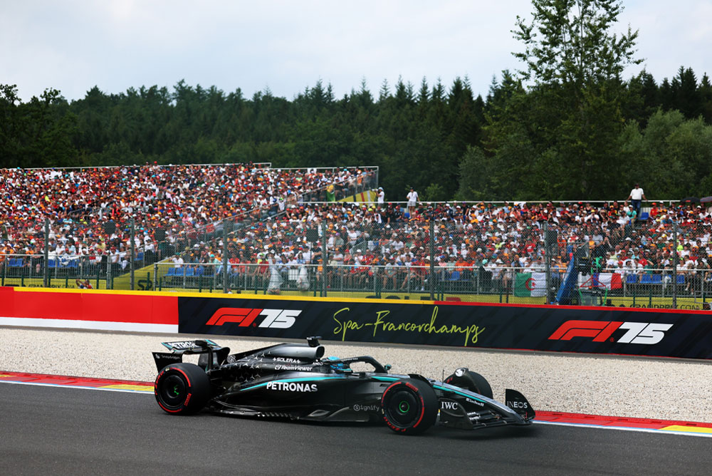 Ein Mercedes-AMG PETRONAS Formel-1-Auto fährt auf dem Circuit Spa-Francorchamps. Im Hintergrund sieht man volle Tribünen mit Fans und internationalen Flaggen. Auf der Streckenbegrenzung ist das Spa-Francorchamps-Logo zu sehen.
