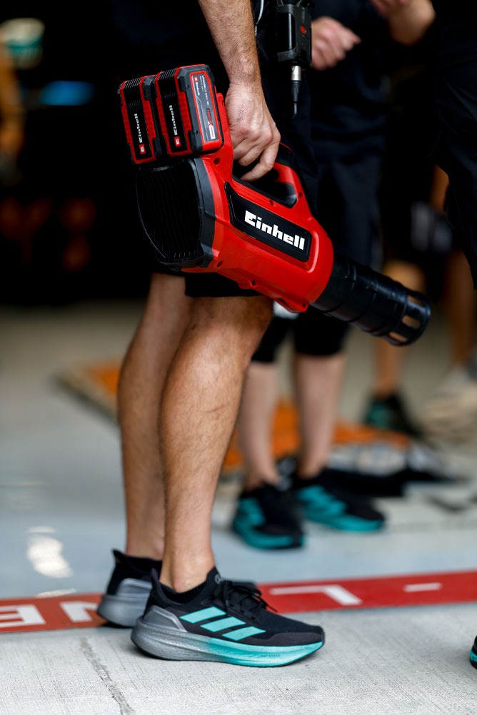 Ein Teammitglied hält einen roten Einhell Akkubläser mit zwei Power X-Change Akkus. Die Szene spielt sich im Boxenbereich bei einem Formel-1-Rennen ab.