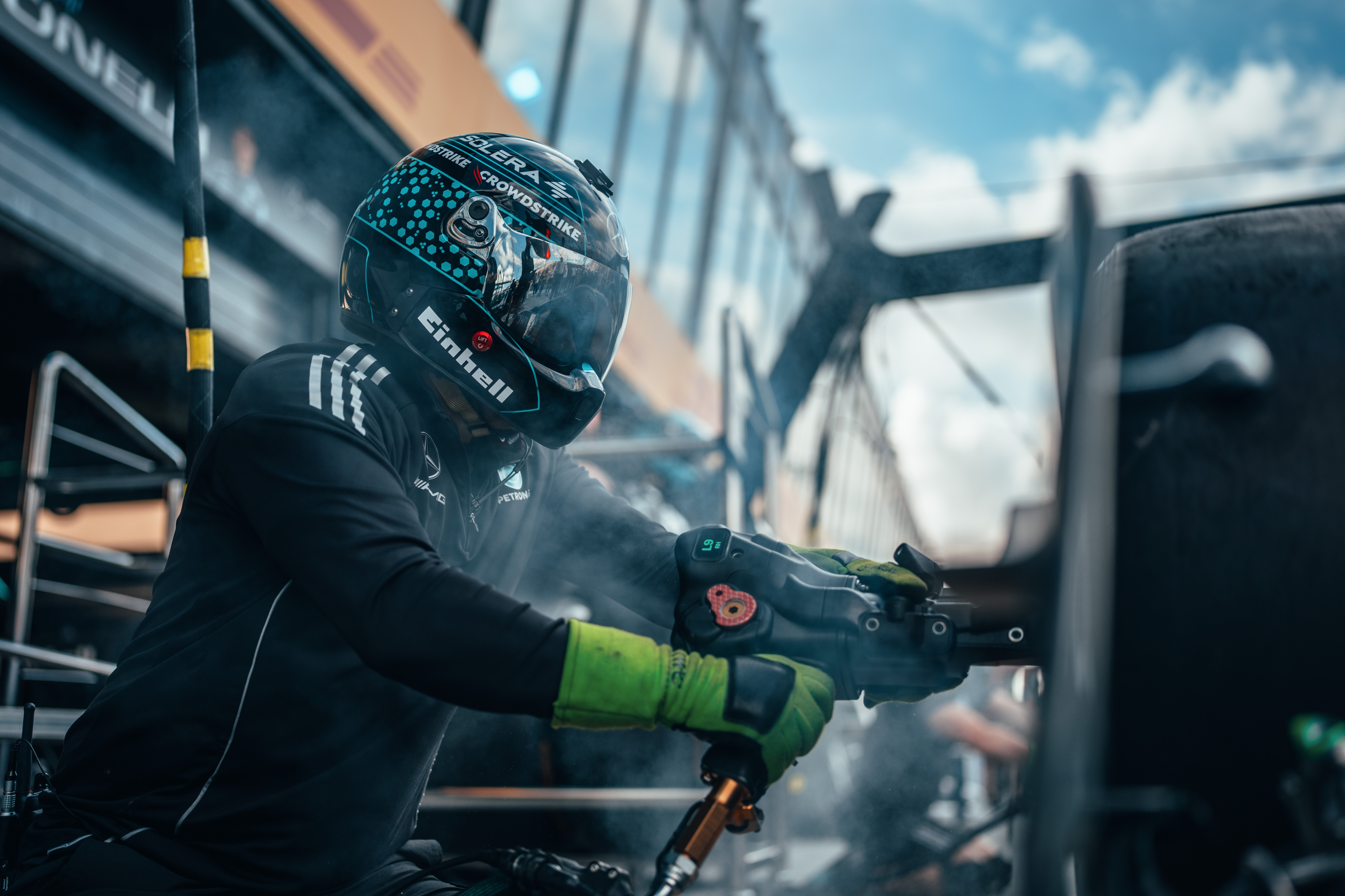 Ein Mitglied der Formel-1-Boxencrew trägt einen Helm und Handschuhe und bedient während eines Boxenstopps eine Druckluft-Radpistole, umgeben von Rauch und Ausrüstung.