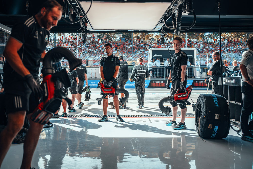 Mechaniker des Mercedes-AMG PETRONAS Formel-1-Teams bereiten sich in der Garage mit kabellosen Einhell-Gebläsen auf einen Boxenstopp vor. Im Hintergrund sind die vollbesetzten Tribünen zu sehen.
