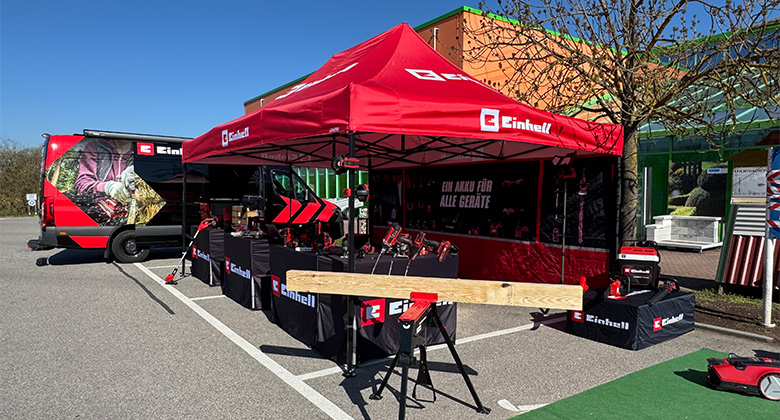 A red Einhell promotional tent with the slogan “One battery for all devices” on a parking lot, surrounded by tools, equipment, and an Einhell van.