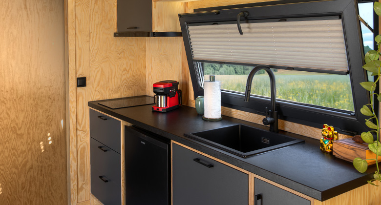 Küchenzeile im Tiny House mit schwarzer Arbeitsplatte, dunklen Unterschränken, Kaffeemaschine und Blick aus dem Fenster.