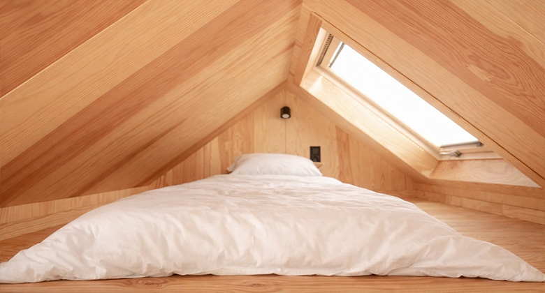 Schlafplatz auf einer Empore unter einer Dachschräge aus hellem Holz mit weißer Matratze und einem Dachfenster.