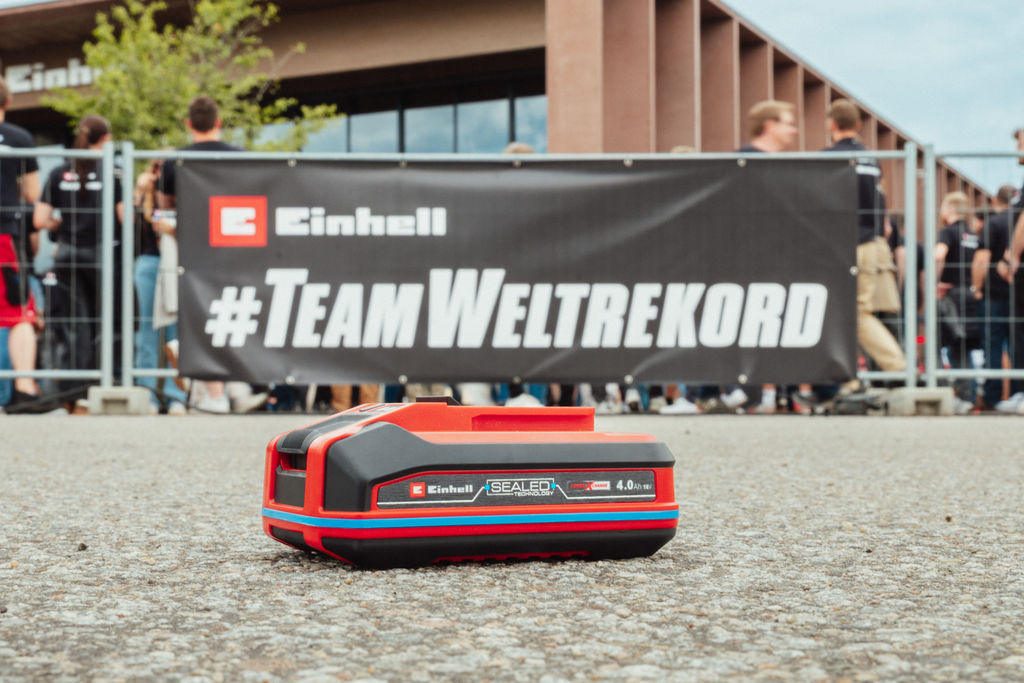 Ein Einhell SEALED Akku Power X-Change 4,0 Ah liegt auf Asphalt, im Hintergrund ein Banner mit „#TeamWeltrekord“.