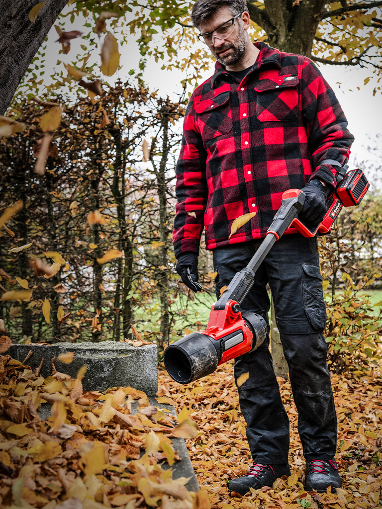 Ein Mann bläst mit einem Einhell Akku-Laubbläser Herbstlaub vor einem Baumstamm weg.