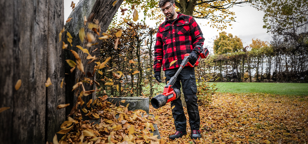 Ein Mann bläst mit einem Einhell Akku-Laubbläser Herbstlaub vor einem Baumstamm weg.