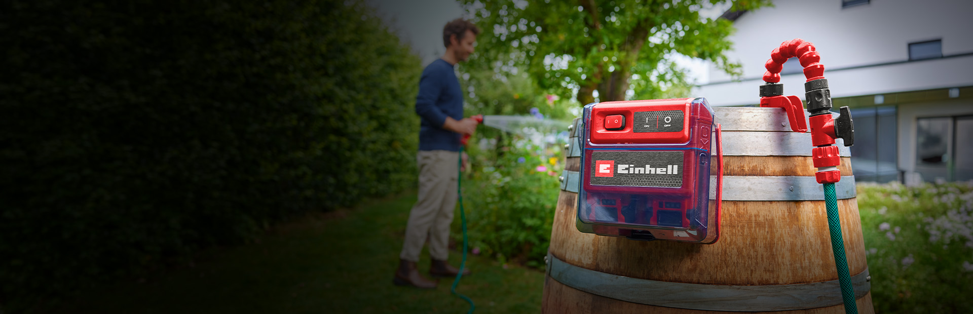 Einhell Akku-Klarwasserpumpe an einem Holzfass im Garten, während eine Person im Hintergrund Pflanzen bewässert.